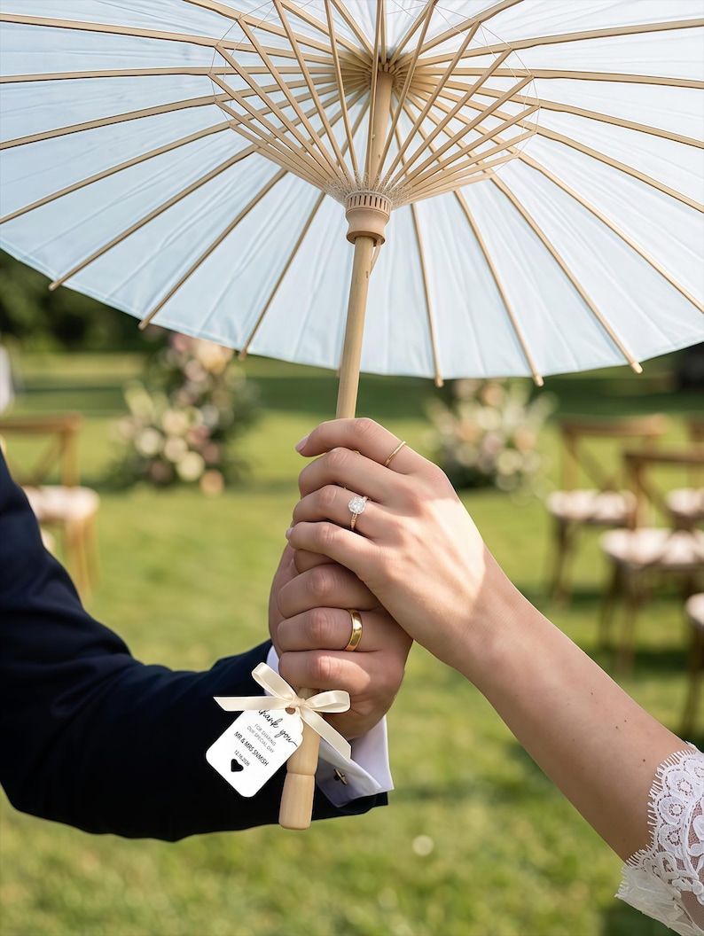 K&ouml;nnte beinhalten: Ein hellblauer Papiersonnenschirm mit Holzgriff und Holzstreben, an dem ein kleines wei&szlig;es Schild mit einem Band befestigt ist. Auf dem Schild steht: Thank you for sharing our special day, Mr & Mrs Samish, 12.16.2026, mit einem schwarzen Herz.