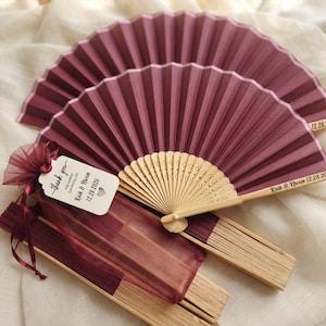 May include: Two deep burgundy folding fans with light wood ribs. One fan is open, displaying the text "Kuik & Nweio 12.28.2026" on the wood. A white tag attached to a burgundy organza bag reads "thank you FOR SHARING OUR SPECIAL DAY Kuik & Nweio 12.28.2026".