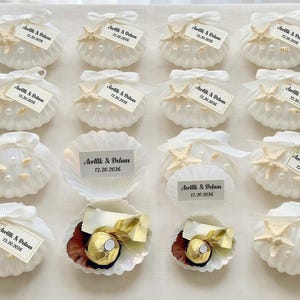 May include: White seashell-shaped favors, adorned with starfish, pearls, and ribbons. Each shell holds a tag with text and the date 12.20.2026. Some shells are open, revealing a chocolate candy.