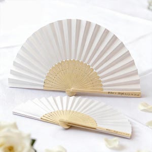 May include: Two white paper fans with natural wood handles. The fans are open, displaying their pleated design. The handles are engraved with text. The fans are likely for a wedding or special event.