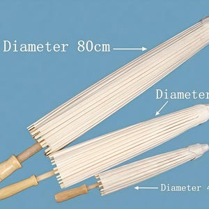 K&ouml;nnte beinhalten: Drei geschlossene wei&szlig;e Papierschirme mit hellen Holzgriffen, diagonal angeordnet. Die Schirme sind mit Durchmessern von 80 cm, 60 cm und 40 cm beschriftet. Das Design zeichnet sich durch eine gerippte Schirmstruktur und einen Holzgriff aus.