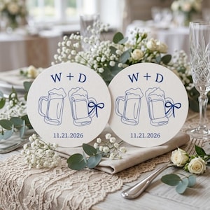 Könnte beinhalten: Zwei runde Untersetzer mit Bierkrug-Design, marineblauem Text "W + D" und Datum "11.21.2026". Sie liegen auf einem Tisch mit Blumenschmuck und Spitzentischdecke, passend für eine Hochzeitsdekoration.