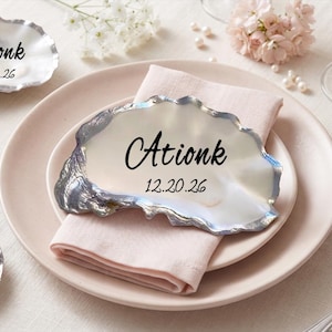 Puede incluir: Una colección de conchas de ostra con bordes pintados en plata, cada una con texto impreso en negro que incluye nombres como Ationk o Dtiomka y la fecha 12.20.26. Las conchas están dispuestas como tarjetas de lugar sobre un plato y mesa.