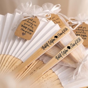 Op de afbeelding: Een verzameling gepersonaliseerde bamboeventilatoren en labels, perfect voor een afstudeerfeest. De ventilatoren zijn lichtbruin en wit, met de tekst "Rokldf Cettylmo Class of 2026" erop gedrukt. Elke ventilator is versierd met een label.