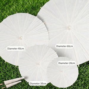 K&ouml;nnte beinhalten: Vier wei&szlig;e Papierschirme mit sichtbaren Holzrippen in verschiedenen Gr&ouml;&szlig;en. Etiketten geben Durchmesser von 20cm, 30cm, 40cm und 60cm an. Zwei weitere geschlossene, gefaltete wei&szlig;e Schirme sind unten links in der Anordnung platziert.
