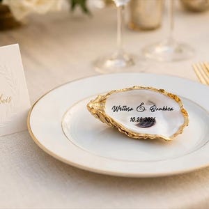 Könnte beinhalten: Hochzeitstafel. Ein weißer Teller mit einer goldfarbenen Austernschale mit handgeschriebenem Text. Eine goldene Gabel und ein Messer befinden sich rechts. Eine Platzkarte mit geprägtem Text befindet sich links.