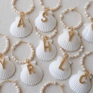 May include: A selection of personalised wine charms. Each charm features a white seashell with a gold initial, suspended from a gold hoop with small, white beads. The charms are arranged on a neutral background.