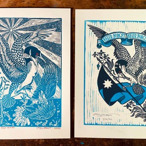 May include: Two blue and white linocut prints. One features a bird with outstretched wings, the other an eagle holding a flag with the text "Never Forget, Never Forgive". Both prints have a hand-carved aesthetic.