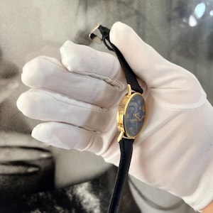 Puede incluir: Un reloj dorado con una esfera azul oscuro con un dise&ntilde;o celestial. El reloj tiene una correa de cuero negro y una caja dorada. El reloj est&aacute; sujeto por una mano enguantada.