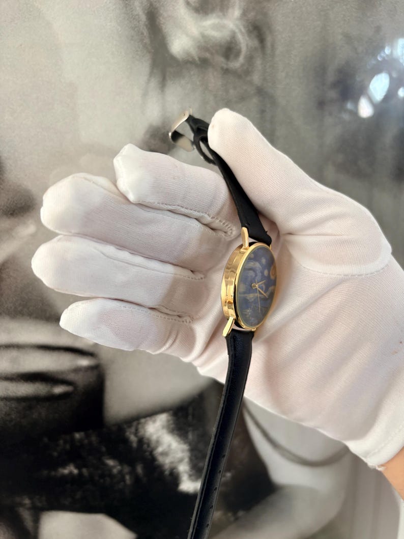 Puede incluir: Un reloj dorado con una esfera azul oscuro con un dise&ntilde;o celestial. El reloj tiene una correa de cuero negro y una caja dorada. El reloj est&aacute; sujeto por una mano enguantada.