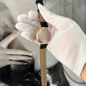 Puede incluir: Un reloj de pulsera dorado con una correa de cuero negro y una parte inferior marr&oacute;n claro. El reloj tiene una esfera redonda y un dise&ntilde;o cl&aacute;sico. El reloj es sostenido por una mano enguantada.