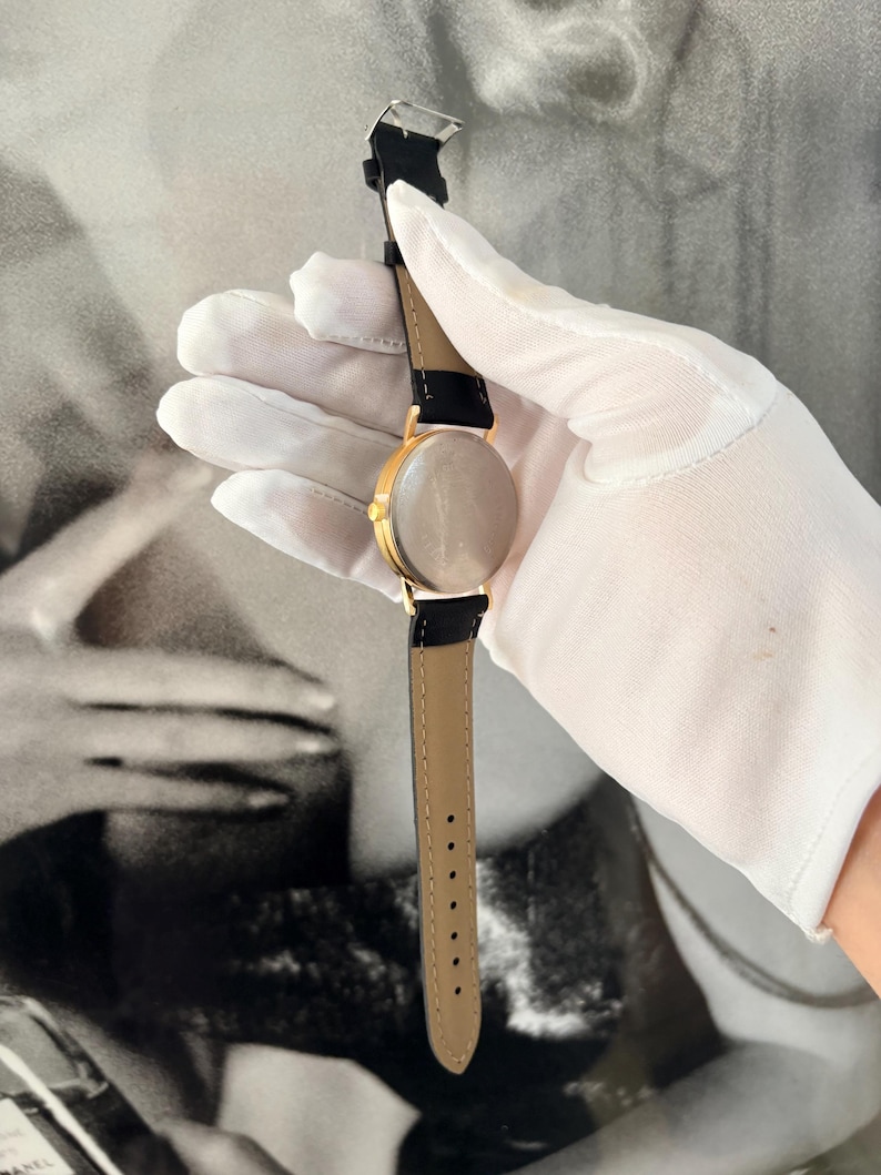 Puede incluir: Un reloj de pulsera dorado con una correa de cuero negro y una parte inferior marr&oacute;n claro. El reloj tiene una esfera redonda y un dise&ntilde;o cl&aacute;sico. El reloj es sostenido por una mano enguantada.