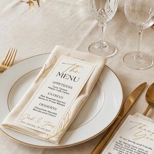 Puede incluir: Un plato llano blanco con borde dorado, cubierto con una servilleta color crema plegada, un menú impreso y una tarjeta de agradecimiento. El menú enumera aperitivos, platos principales y postres. Cubiertos dorados, copas de cristal y flores.