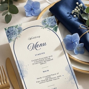 May include: A wedding menu card with a floral design featuring blue hydrangeas. The menu lists starter, main, and dessert options. Gold cutlery and a navy blue napkin are also visible, along with a white rose and champagne glasses.