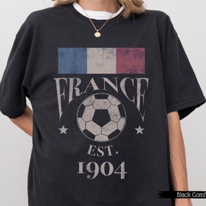 以下が含まれることがあります： フランス国旗のグラフィック、ヴィンテージフォントの「FRANCE」の文字、サッカーボール、そして「EST. 1904」のテキストが特徴の黒いTシャツ。クルーネックと半袖です。