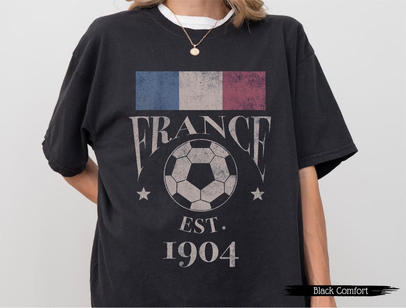 以下が含まれることがあります： フランス国旗のグラフィック、ヴィンテージフォントの「FRANCE」の文字、サッカーボール、そして「EST. 1904」のテキストが特徴の黒いTシャツ。クルーネックと半袖です。
