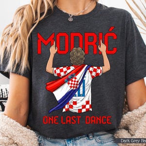 May include: Dark grey heather t-shirt featuring the name "MODRIĆ" in red, with a graphic of a person wearing a Croatian flag. The text "ONE LAST DANCE" is below the graphic. The shirt is a dark grey color.