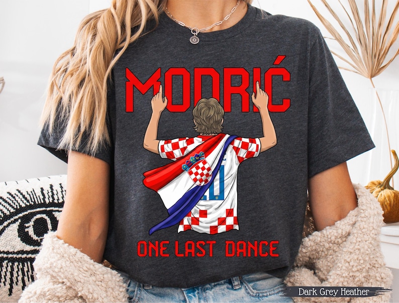 May include: Dark grey heather t-shirt featuring the name "MODRIĆ" in red, with a graphic of a person wearing a Croatian flag. The text "ONE LAST DANCE" is below the graphic. The shirt is a dark grey color.