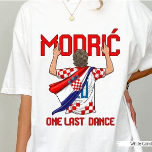 May include: White t-shirt featuring a graphic of a soccer player wearing a Croatian flag. The word "MODRIĆ" is in red block letters above the player, with the text "ONE LAST DANCE" below. The shirt is labeled "White Comfort".