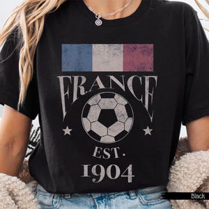 以下が含まれることがあります： フランスの国旗、"FRANCE"の文字、サッカーボール、"EST. 1904"の文字がプリントされた黒いTシャツ。 ヴィンテージ感のあるデザイン。 カジュアルウェアアイテム。