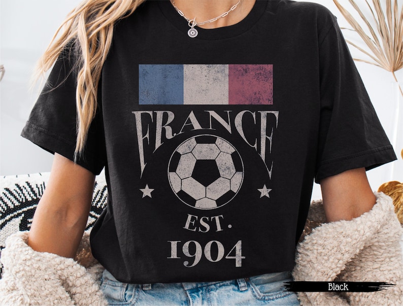 以下が含まれることがあります： フランスの国旗、"FRANCE"の文字、サッカーボール、"EST. 1904"の文字がプリントされた黒いTシャツ。 ヴィンテージ感のあるデザイン。 カジュアルウェアアイテム。