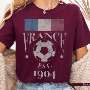 以下が含まれることがあります： フランス国旗のグラフィックが施された、色あせたマルーン色のTシャツ。「FRANCE」の文字はヴィンテージフォントで、サッカーボールと「EST. 1904」の文字がプリントされています。サッカーボールの両側に2つの星があります。