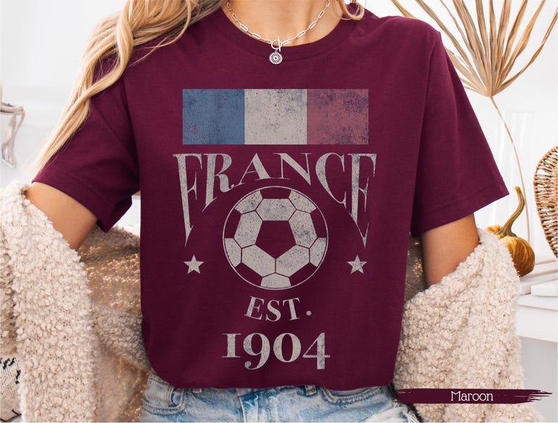 以下が含まれることがあります： フランス国旗のグラフィックが施された、色あせたマルーン色のTシャツ。「FRANCE」の文字はヴィンテージフォントで、サッカーボールと「EST. 1904」の文字がプリントされています。サッカーボールの両側に2つの星があります。