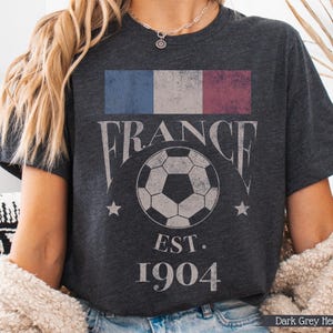 以下が含まれることがあります： フランスの国旗、"FRANCE"の文字、サッカーボール、2つの星、"EST. 1904"の文字がプリントされた、色あせたグラフィックのダークグレーのヘザーTシャツ。クルーネックと半袖です。