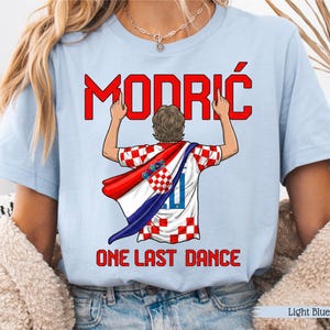 May include: Light blue t-shirt featuring the name "MODRIĆ" in red above an illustration of a soccer player wearing a Croatian flag. The text "ONE LAST DANCE" is below the illustration. The shirt is light blue.