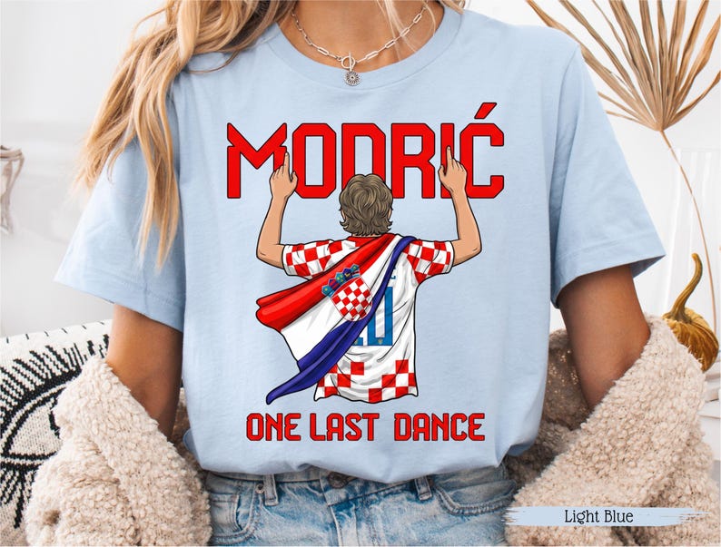May include: Light blue t-shirt featuring the name "MODRIĆ" in red above an illustration of a soccer player wearing a Croatian flag. The text "ONE LAST DANCE" is below the illustration. The shirt is light blue.