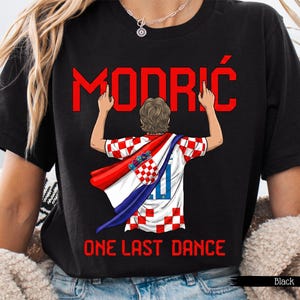 May include: Black t-shirt featuring a graphic of a soccer player in a Croatian jersey with the name "MODRIĆ" in red above. The text "ONE LAST DANCE" is below the graphic. The player is wearing a red and white cape.