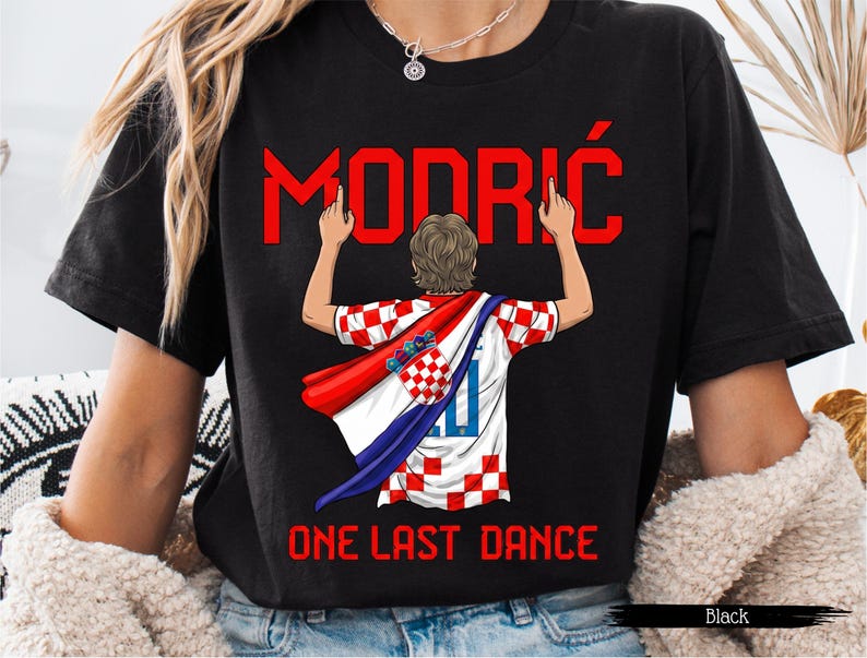 May include: Black t-shirt featuring a graphic of a soccer player in a Croatian jersey with the name "MODRIĆ" in red above. The text "ONE LAST DANCE" is below the graphic. The player is wearing a red and white cape.