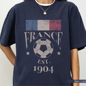以下が含まれることがあります： フランス国旗のデザイン、"FRANCE"の文字、サッカーボールのグラフィックがプリントされたネイビーブルーのTシャツ。ボールの下には"EST. 1904"の文字。快適なフィット感のシャツです。