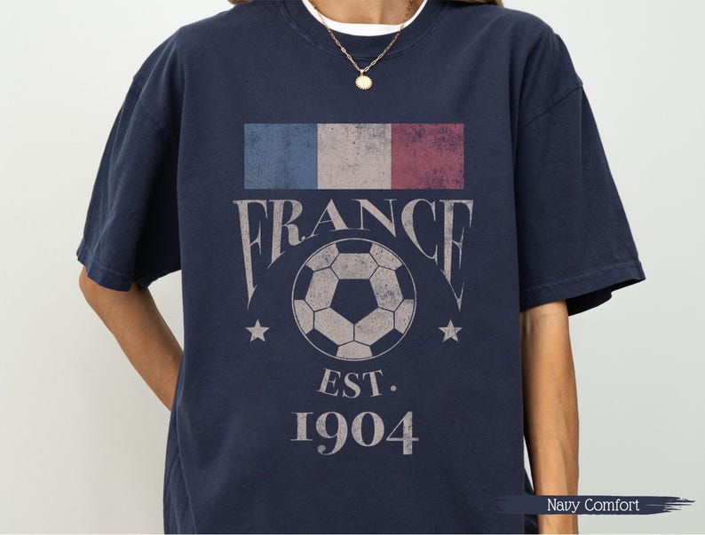 以下が含まれることがあります： フランス国旗のデザイン、"FRANCE"の文字、サッカーボールのグラフィックがプリントされたネイビーブルーのTシャツ。ボールの下には"EST. 1904"の文字。快適なフィット感のシャツです。
