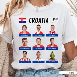 以下が含まれることがあります： クロアチアの国旗と「-CROATIA- SQUAD LIST」の文字が入った白いTシャツ。サッカー選手とグリンチの画像が表示されています。Tシャツの下部には「White」の文字があります。