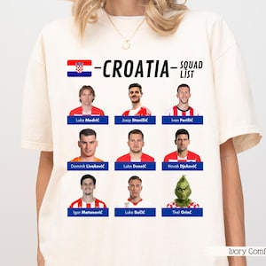 以下が含まれることがあります： 「CROATIA」の文字とクロアチア国旗のグラフィックがプリントされた、アイボリーコンフォートのクリーム色のTシャツ。選手の写真と名前が記載されたチームリストがあり、下部にはグリンチのグラフィックがあります。