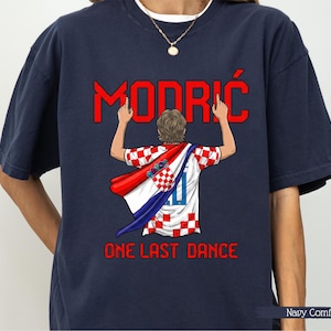 May include: Navy blue t-shirt featuring a graphic of a soccer player with a Croatian flag cape. The word "MODRIĆ" is in red above the player, and "ONE LAST DANCE" is below. The shirt is labeled "Navy Comfort".