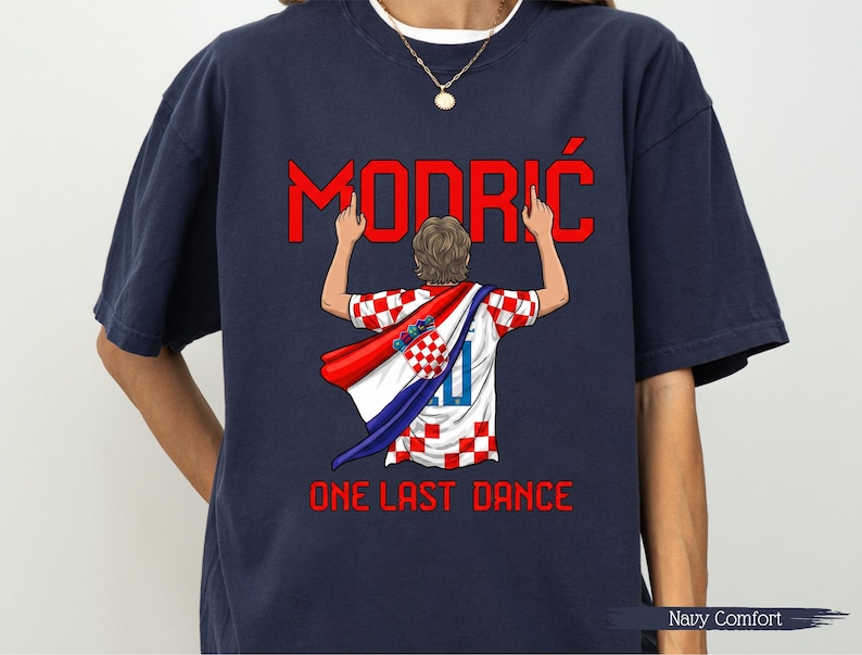 May include: Navy blue t-shirt featuring a graphic of a soccer player with a Croatian flag cape. The word "MODRIĆ" is in red above the player, and "ONE LAST DANCE" is below. The shirt is labeled "Navy Comfort".