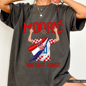 May include: Dark grey t-shirt featuring the name "Modrić" in red, above an illustration of a soccer player wearing a Croatian flag. The text "ONE LAST DANCE" is below the illustration. The shirt has a vintage, washed look.