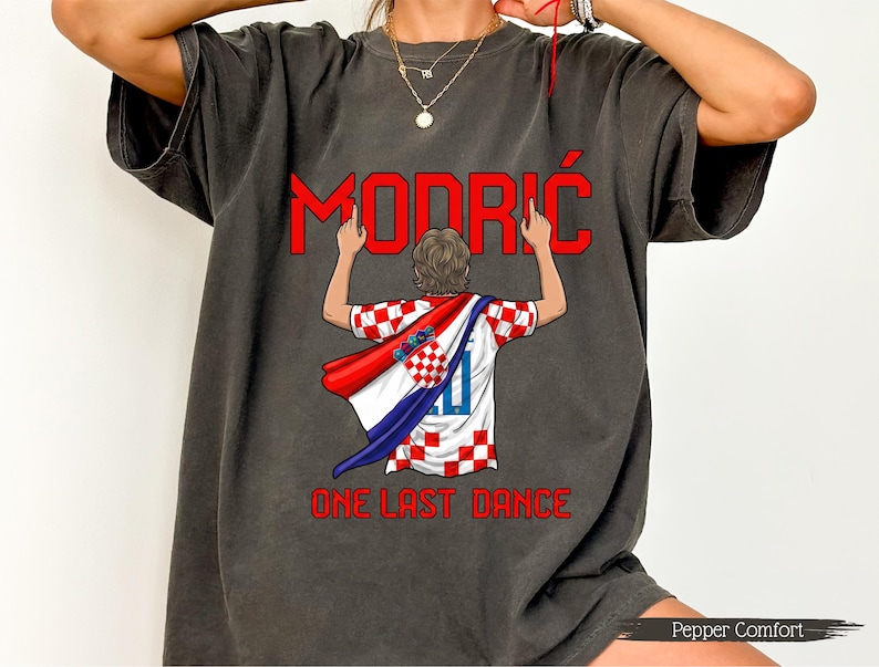 May include: Dark grey t-shirt featuring the name "Modrić" in red, above an illustration of a soccer player wearing a Croatian flag. The text "ONE LAST DANCE" is below the illustration. The shirt has a vintage, washed look.