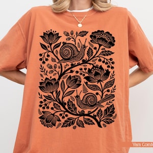 May include: Orange t-shirt with a black floral and snail design. The graphic features two snails among flowers, leaves, and berries. The shirt has short sleeves and a relaxed fit. The text "Yam Comfort" is at the bottom right.