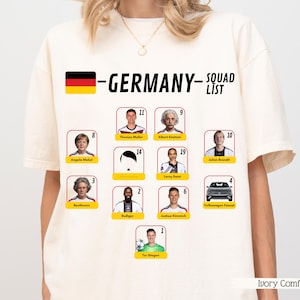 Könnte beinhalten: Ivory Comfort T-Shirt mit der deutschen Flagge und dem Wort "GERMANY" über einer "SQUAD LIST". Die Liste enthält Porträts berühmter Deutscher und ein Auto. Das T-Shirt ist elfenbeinfarben.