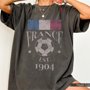 以下が含まれることがあります： フランスの国旗、"FRANCE"の文字、サッカーボール、"EST. 1904"の文字がプリントされたダークグレーのTシャツ。半袖でゆったりとしたフィット感。柔らかい素材で作られています。