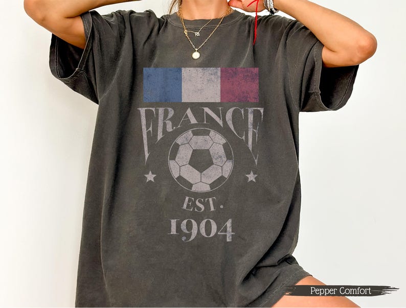 以下が含まれることがあります： フランスの国旗、"FRANCE"の文字、サッカーボール、"EST. 1904"の文字がプリントされたダークグレーのTシャツ。半袖でゆったりとしたフィット感。柔らかい素材で作られています。
