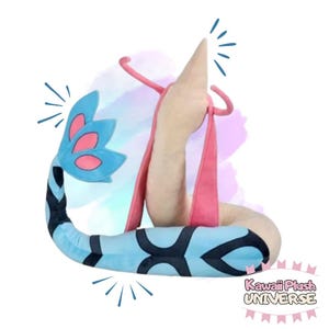 May include: A plush toy in the shape of a stylized creature, featuring a light blue body with black markings, a pink strap, and a beige head. The tail has blue and pink accents. The text "Kawaii Plush UNIVERSE" is visible.