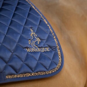 Puede incluir: Un salvacruces acolchado azul marino con un logotipo dorado bordado de un caballo y una corona, y la palabra "MONARQUE". La almohadilla tiene un borde con estampado de leopardo, diseñado para uso ecuestre.