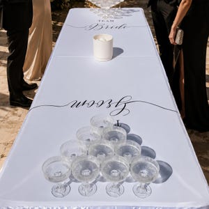 Op de afbeelding: Een wit rechthoekig tafelkleed met de zwarte tekst Team Bride en Groom voor een spel. Het oppervlak bevat een witte cilindrische beker en tien heldere, geribbelde glazen coupes die in een driehoeksvorm op de tafel zijn geplaatst.