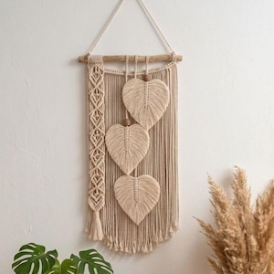 May include: A cream-colored macrame wall hanging suspended from a wooden branch. It features a vertical braided panel on the left and three heart-shaped leaf motifs hanging in the center, each accented with a small wooden bead at the top.