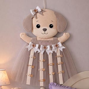May include: A wall-hanging felt dog organiser featuring the name Kamila embroidered on the bodice. The dog has tan felt ears, a white bow on its head, and a beige tulle skirt with vertical ribbons holding several gold-toned hair clips.