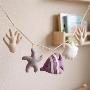 Puede incluir: Una guirnalda de fieltro con motivos marinos que presenta una estrella de mar, un pez, una concha y coral en tonos morado, blanco y beige. La guirnalda está ensartada en una cuerda natural, creando un tema náutico caprichoso.
